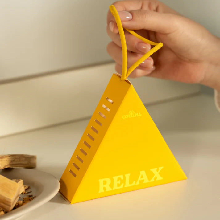 Yellow pyramid-shaped fragrance sachet with 'RELAX' text held by a hand on a neutral background