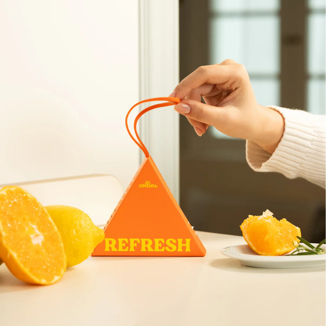 Orange triangular fruity fragrance sachet with a hand holding it next to fruits on a table.