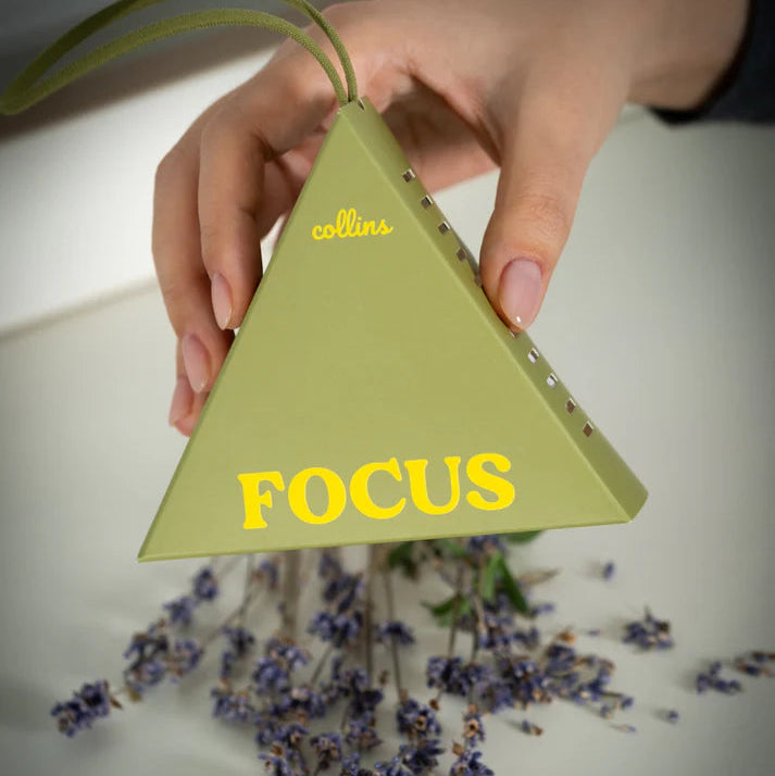 Triangular green fragrance sachet with 'Collins Focus' text held by a hand with lavender flowers.