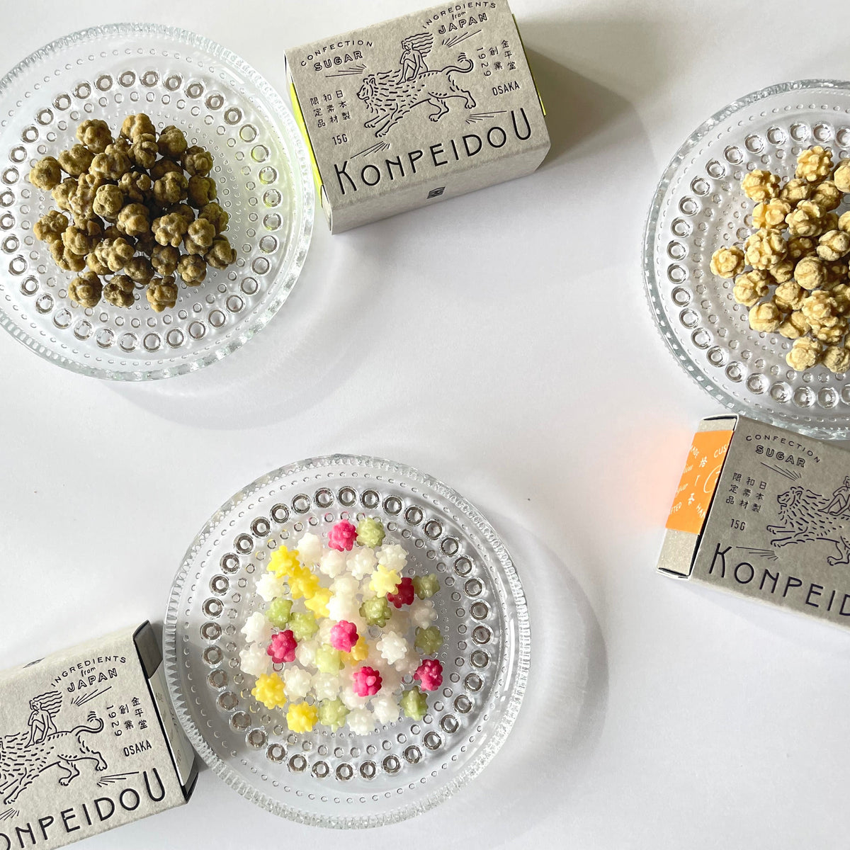 Four small clear glass dishes containing assorted popcorn kernels and candy-like pieces, arranged on a white surface with two visible Konpeidou matchboxes nearby.