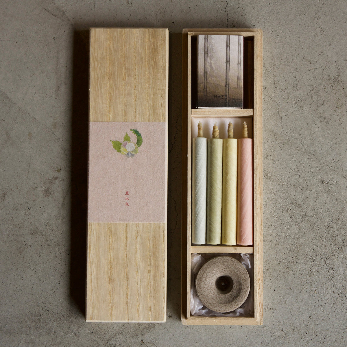 A Japanese-style incense set in a light wood box, featuring a tall rectangular box with a beige paper band and a smaller open compartment holding incense sticks in cream, yellow, and pink, along with a rolled incense burner, all placed on a textured gray concrete surface.