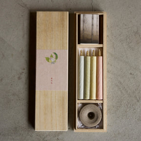 A Japanese-style incense set in a light wood box, featuring a tall rectangular box with a beige paper band and a smaller open compartment holding incense sticks in cream, yellow, and pink, along with a rolled incense burner, all placed on a textured gray concrete surface.
