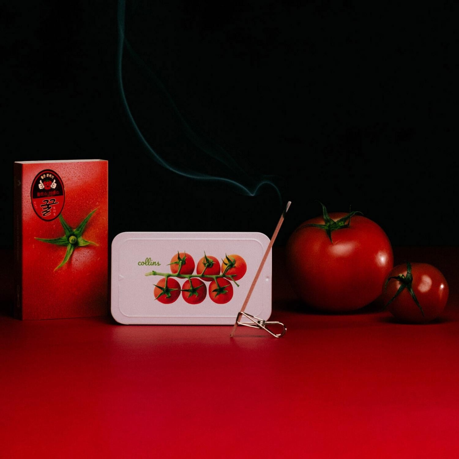 A styled product shot featuring a white rectangular tin with a red cherry tomato illustration, next to a red box with a black circular tomato graphic. A lit incense stick with blue-green smoke rests on a metal stand in front of the tin.