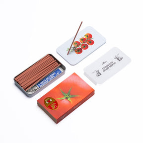 A set of incense sticks in a silver metal tin with a white lid printed with red tomatoes and a single incense stick resting on it. Another incense stick lies on the front orange-red box, which features a green lotus flower and a red circular seal.