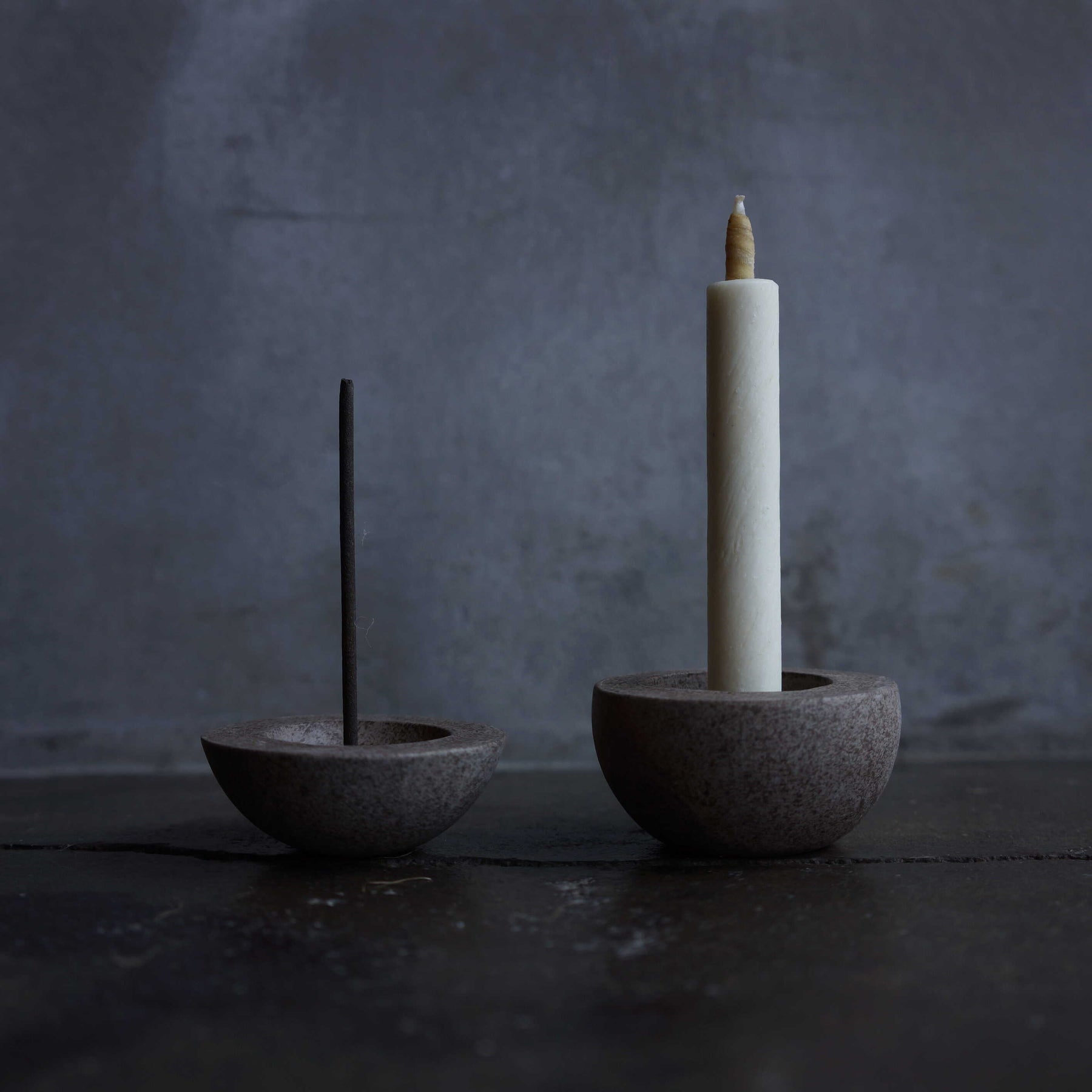 Two stone incense holders with a lit candle and unlit incense stick on a dark background