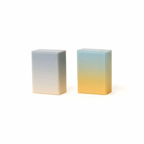 Two rectangular gradient blocks of soap standing upright on a white background, one fading from pale blue-gray to off-white and the other from pale blue to warm yellow.