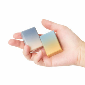 A hand holding two small rectangular gradient items of soap, one transitioning from blue-gray to white, and the other from light blue to warm yellow, against a plain white background.
