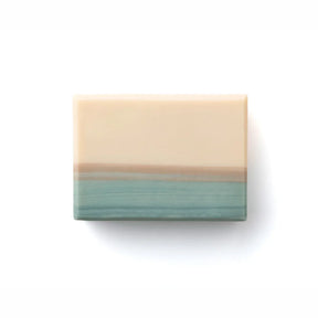 A rectangular bar of soap with a two-tone design, featuring a warm cream-colored top half and a textured teal-green bottom half with a thin tan band in the middle, shown on a plain white background.